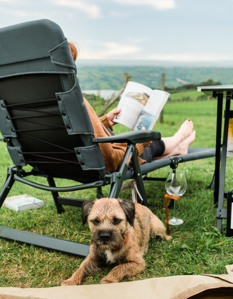 A Woman Sits In A Camping Chair Reading A Travel Bookwith Her Border Terrier By Her Side, Overlooking A View Into The Distance