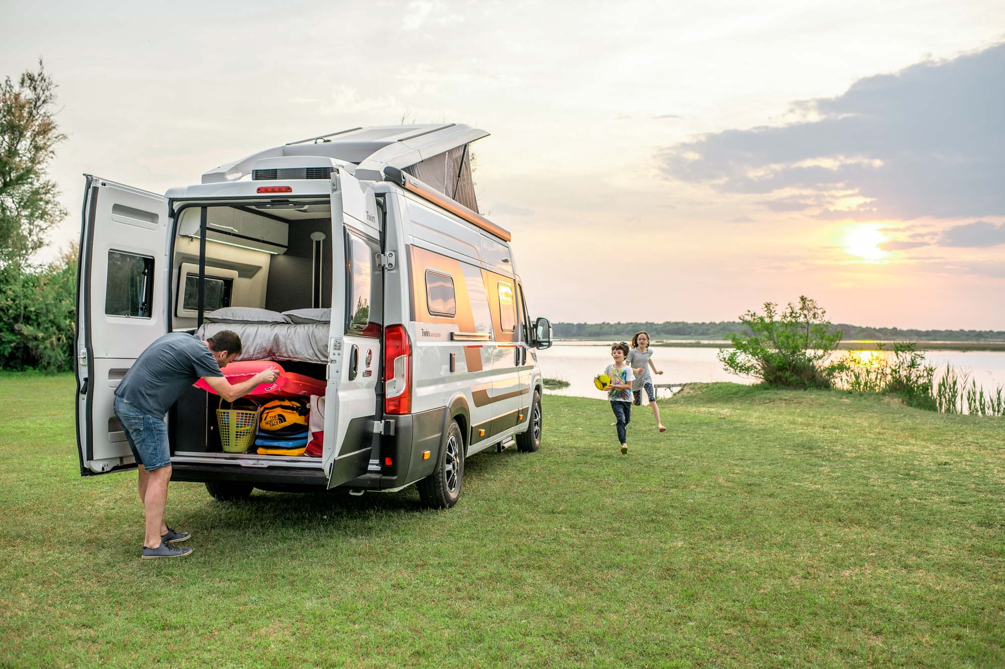 A Man Looks In The Boot Of His Van Conversion Motorhome While Two Children Run Playing Games, With A Lake And Sunset In The Distance